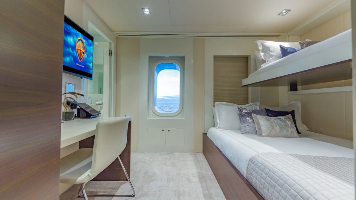 Cozy cabin with a window showcasing an ocean view. The room features a bunk bed, a single bed, a desk with a chair, and a mounted TV on the wall. Soft lighting and neutral tones create a relaxing atmosphere.
