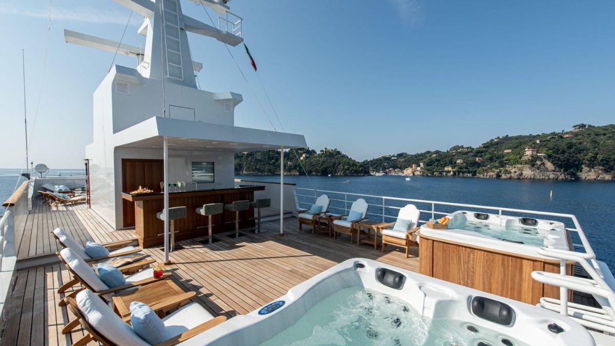 Luxury yacht deck featuring two hot tubs, wooden loungers, and a bar area. The tranquil sea and lush greenery outline the horizon under a clear blue sky.