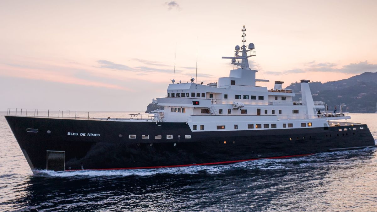 A large, modern yacht named "Bleu de Nîmes" sails on calm waters during sunset, showcasing its sleek black hull and multiple decks. The scene features a colorful sky with hints of orange and pink reflecting on the water.