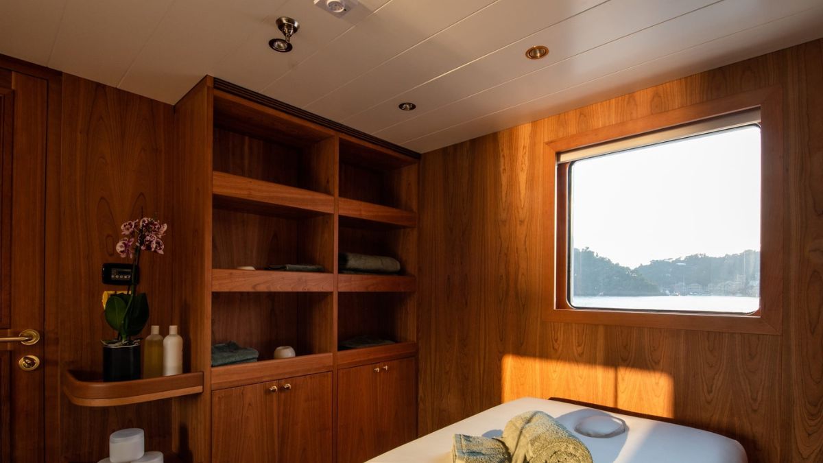 A wooden interior of a luxury cabin with shelves containing rolled towels and toiletries. A large window offers a view of the outdoors, while a massage table is set up in the center, adorned with soft linens.
