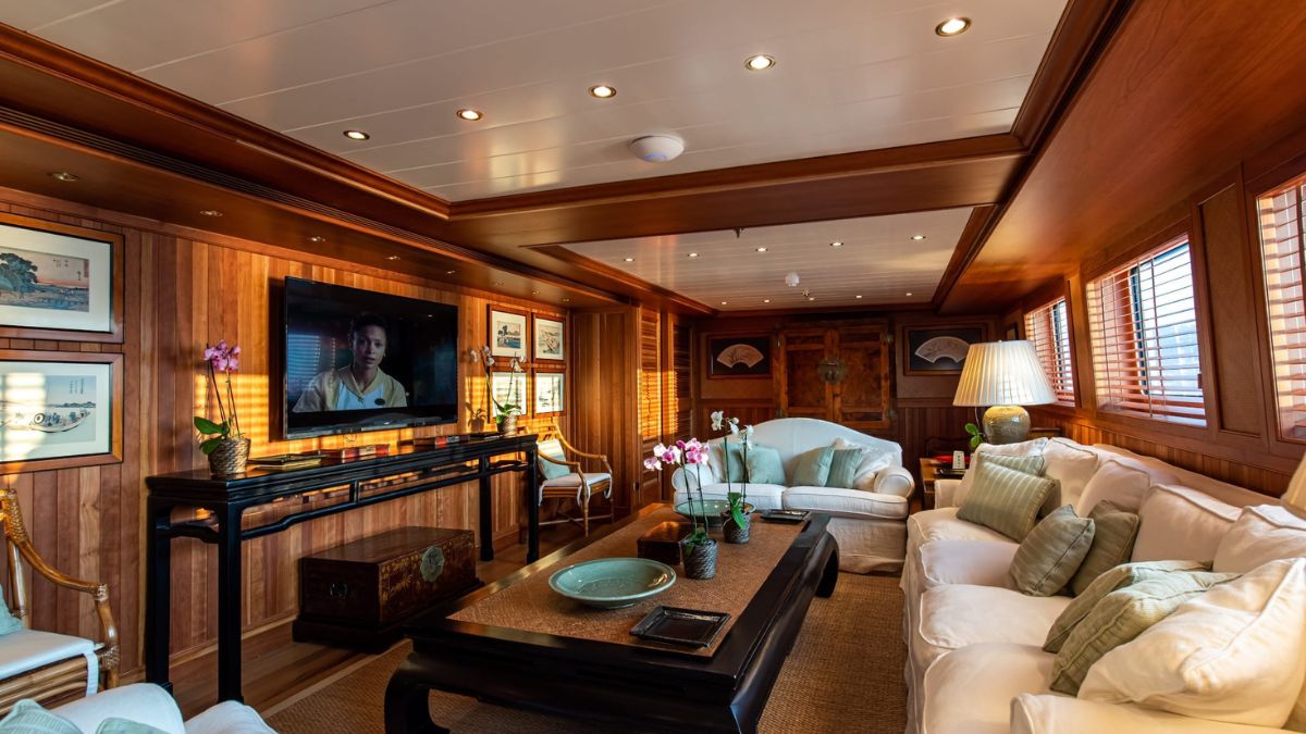 A luxurious living room with wooden paneling, featuring a comfortable seating area with white sofas and decorative pillows. A large television is mounted on the wall, and there are elegant decorations, including framed artwork and a lamp. Natural light filters through the wooden blinds.