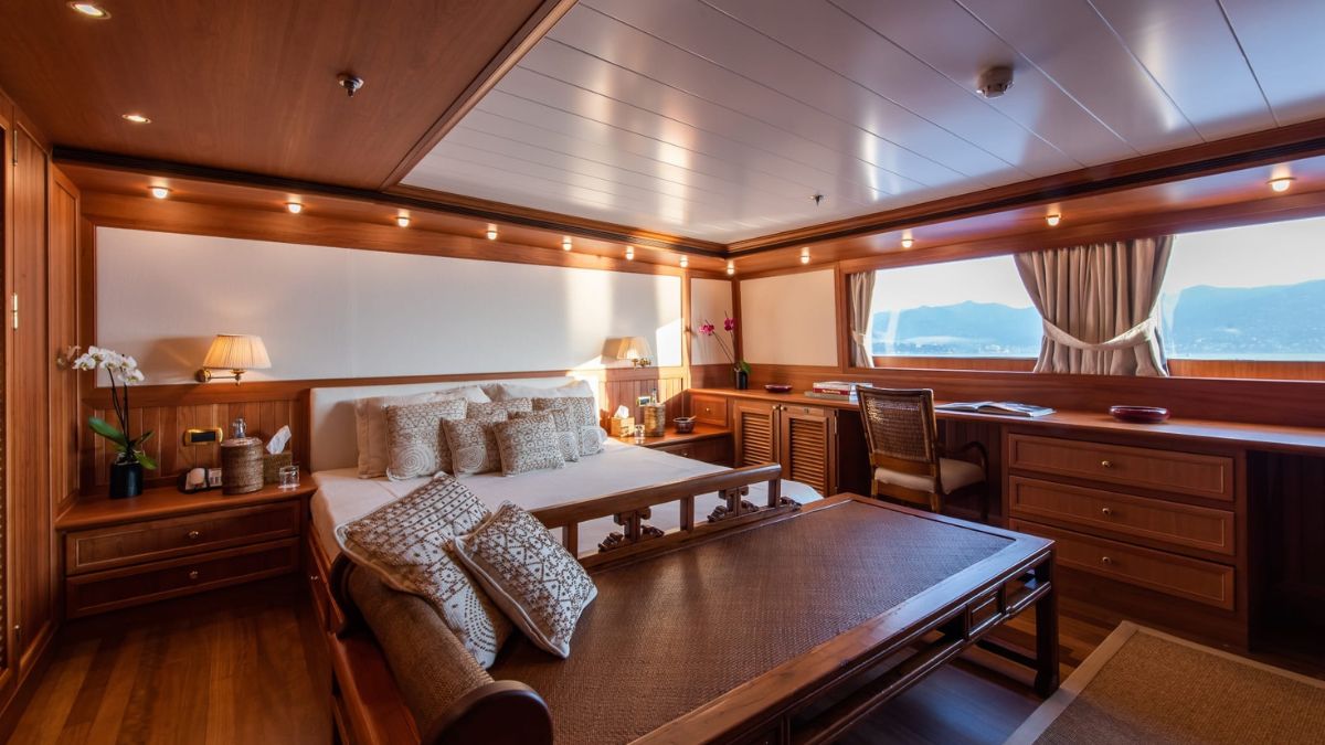 A spacious, elegantly designed bedroom with warm wood paneling. The room features a large bed with decorative pillows, a comfortable seating area, and a stylish desk. Sunlight streams through a large window, illuminating mountains in the background.