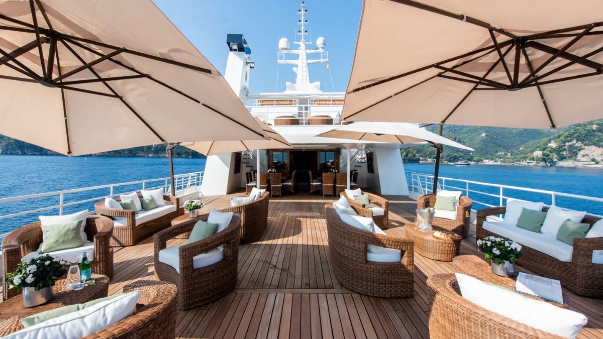 A luxurious yacht deck featuring comfortable wicker seating arrangements, large umbrellas providing shade, and beautiful views of the water and mountains in the background.