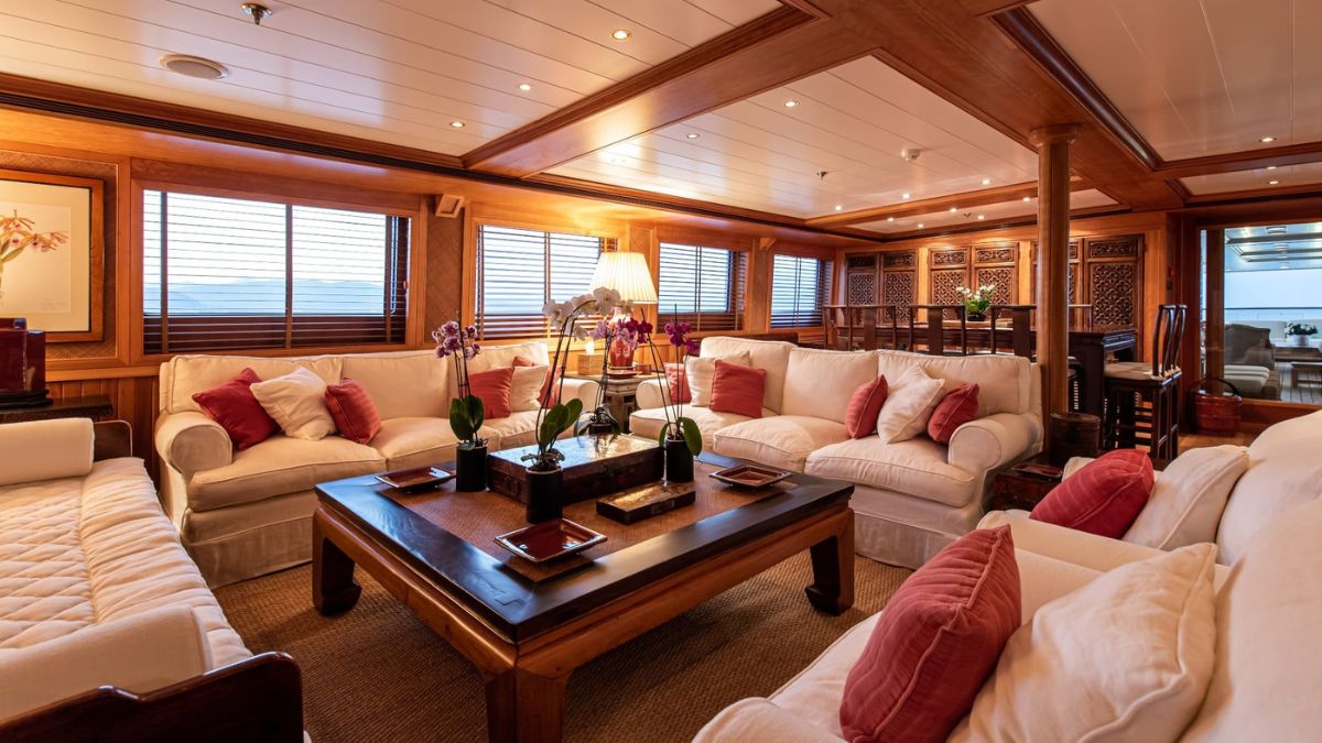 Elegant yacht interior featuring a spacious living area with white sofas adorned with red cushions, a wooden coffee table, and soft lighting. Large windows provide natural light, and decorative plants add a touch of greenery. A dining area is visible in the background, showcasing ornate woodwork.