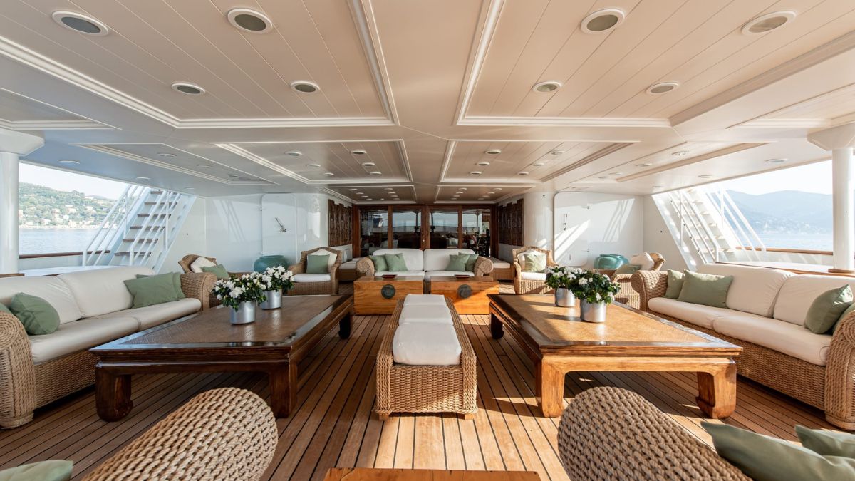 Spacious outdoor lounge area on a luxury yacht, featuring comfortable sofas, wooden coffee tables, and decorative floral arrangements. Bright natural light fills the space, with views of the water visible in the background.