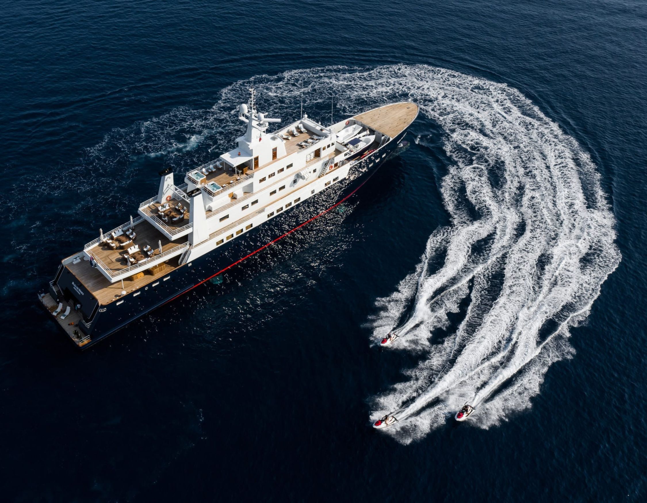 A large luxury yacht sails through deep blue water, trailing two smaller speedboats that create swirling wakes. The yacht features a spacious upper deck with lounge areas.