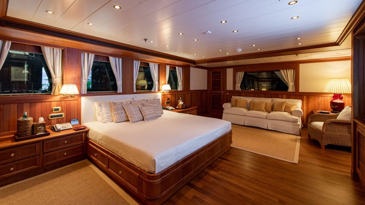 Elegant wooden bedroom featuring a king-size bed with decorative pillows, a plush sofa, warm lighting, and large windows. The floor is covered in a soft rug, and wooden panels adorn the walls, creating a cozy atmosphere.