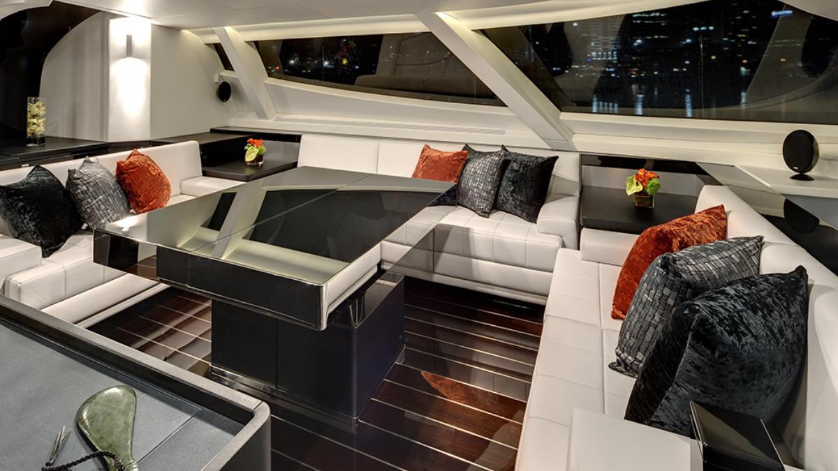 A modern yacht interior featuring a sleek black and white seating area with plush cushions in shades of grey and orange. Large windows provide a view of the city skyline, and a contemporary coffee table sits in the centre.