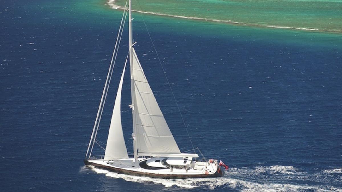 A sleek sailing yacht glides through deep blue waters, with large white sails billowing in the wind. In the background, a vibrant turquoise lagoon contrasts with the ocean.