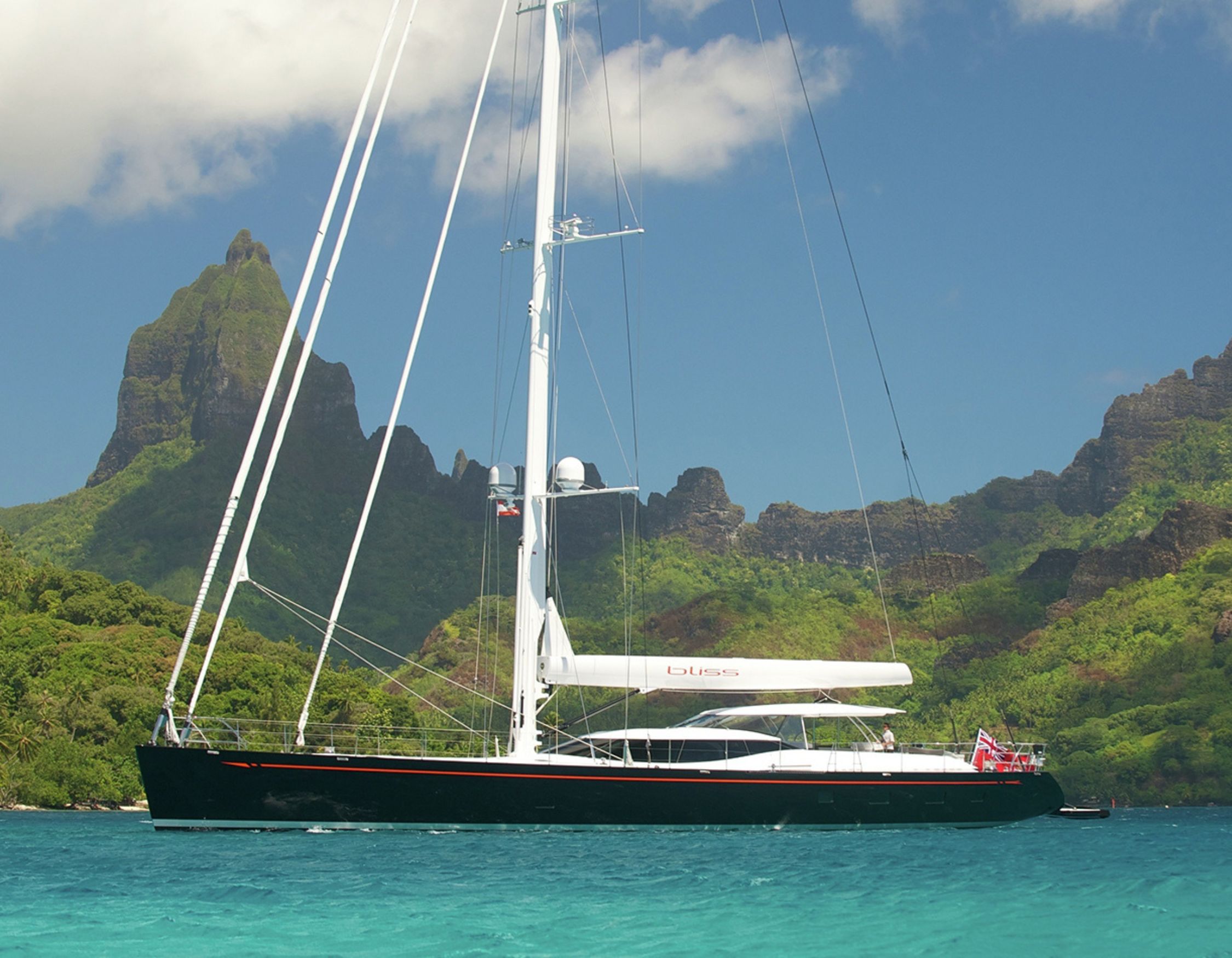 A sleek sailboat anchored in turquoise waters, with a backdrop of lush green mountains and a partly cloudy sky.