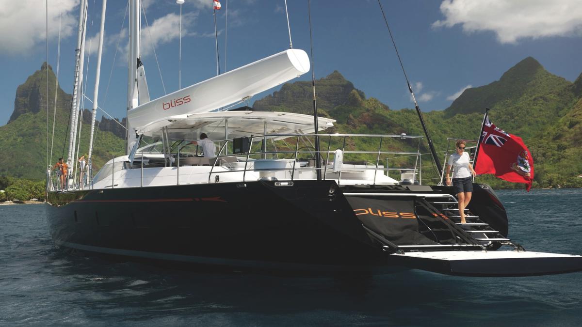 A sleek black yacht named "Bliss" anchored in a calm bay, surrounded by lush green mountains under a partly cloudy sky. A person stands on the boat's deck, and a British flag flutters from the stern.
