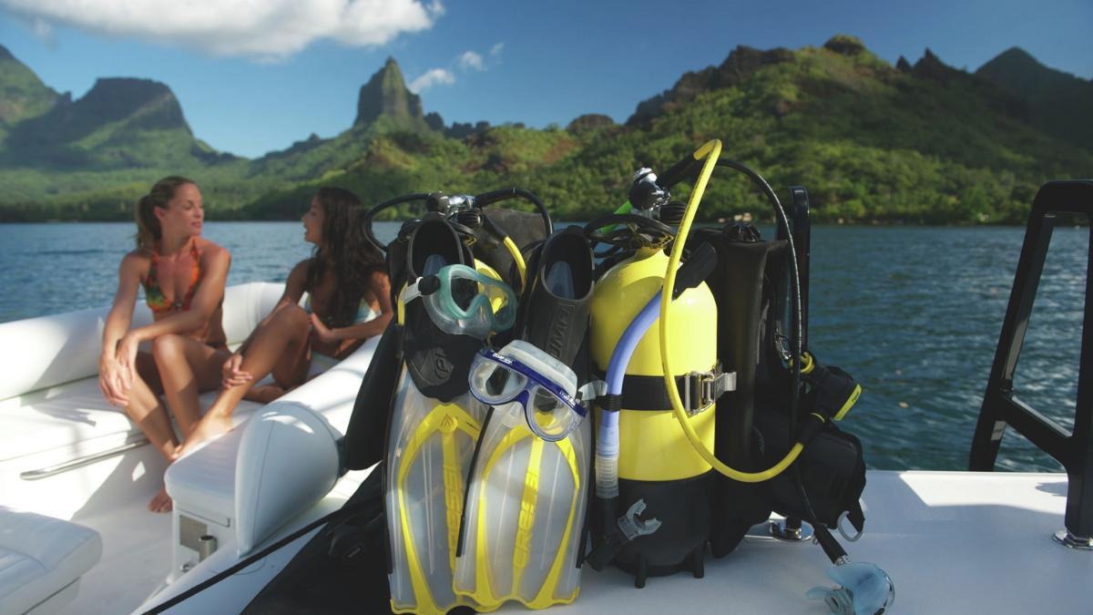 Two women relax on a boat, chatting in front of lush green mountains. Scuba gear is arranged on the boat, including fins, snorkel, and a yellow tank. The scene captures a serene day on the water.
