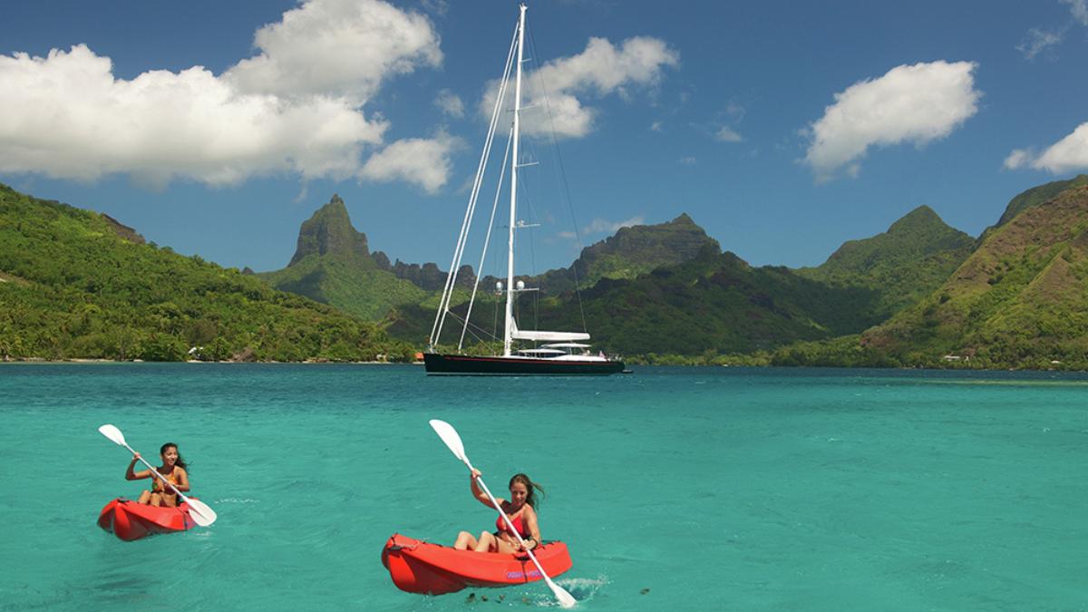 Two people kayaking in bright red kayaks on turquoise water, surrounded by lush green mountains and a large sailing yacht in the background under a blue sky with fluffy white clouds.