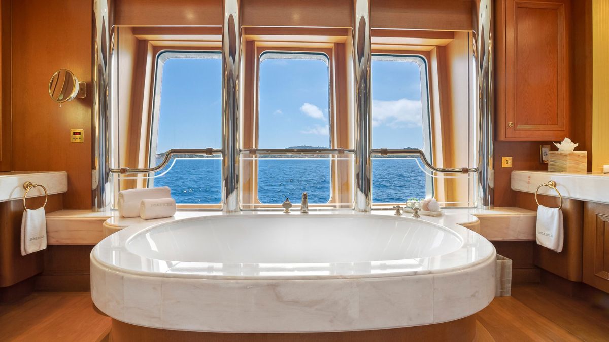 A luxurious bathroom featuring a large white bathtub, wooden cabinetry, and large windows showcasing a view of the ocean and blue sky. Plush towels are neatly arranged nearby.