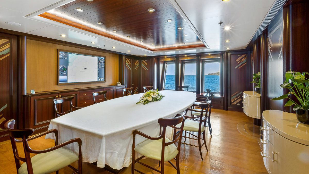 A elegant boardroom with a long, white table surrounded by wooden chairs. Large windows offer a view of the ocean, and a wall-mounted screen displays a map. The room features wood paneling and decorative accents, with plants in the corners.