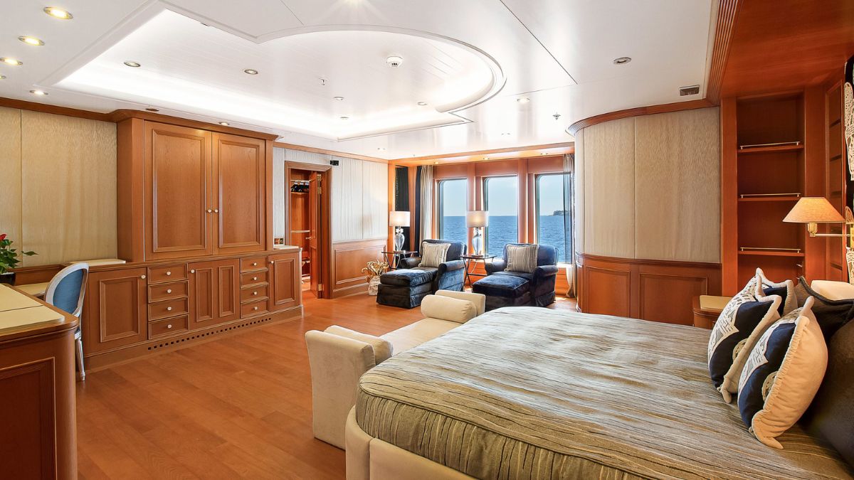 Luxurious yacht interior featuring a spacious bedroom with a large bed, wooden cabinetry, and two armchairs facing panoramic windows overlooking the sea. Natural light enhances the elegant decor.