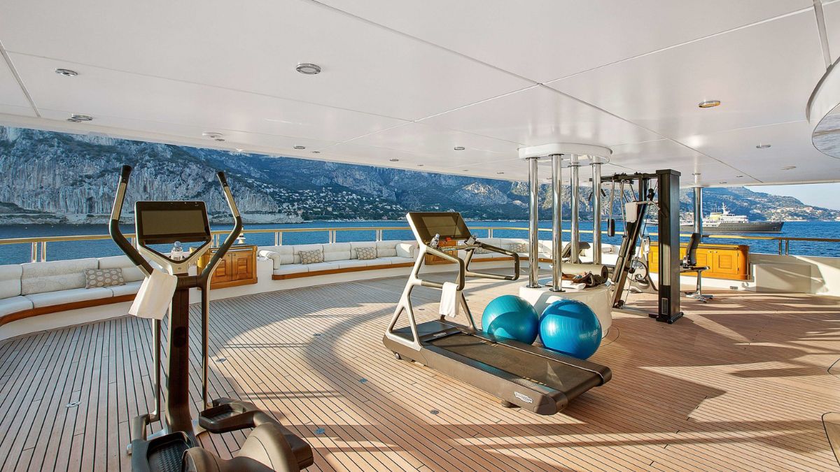 A luxurious gym on a yacht, featuring elliptical machines, treadmills, and exercise balls. Scenic views of mountains and sea are visible in the background.