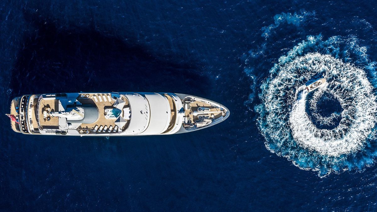 Aerial view of a large yacht anchored in deep blue water, with a smaller boat creating a circular wake nearby. The yacht features a spacious deck with lounge areas.