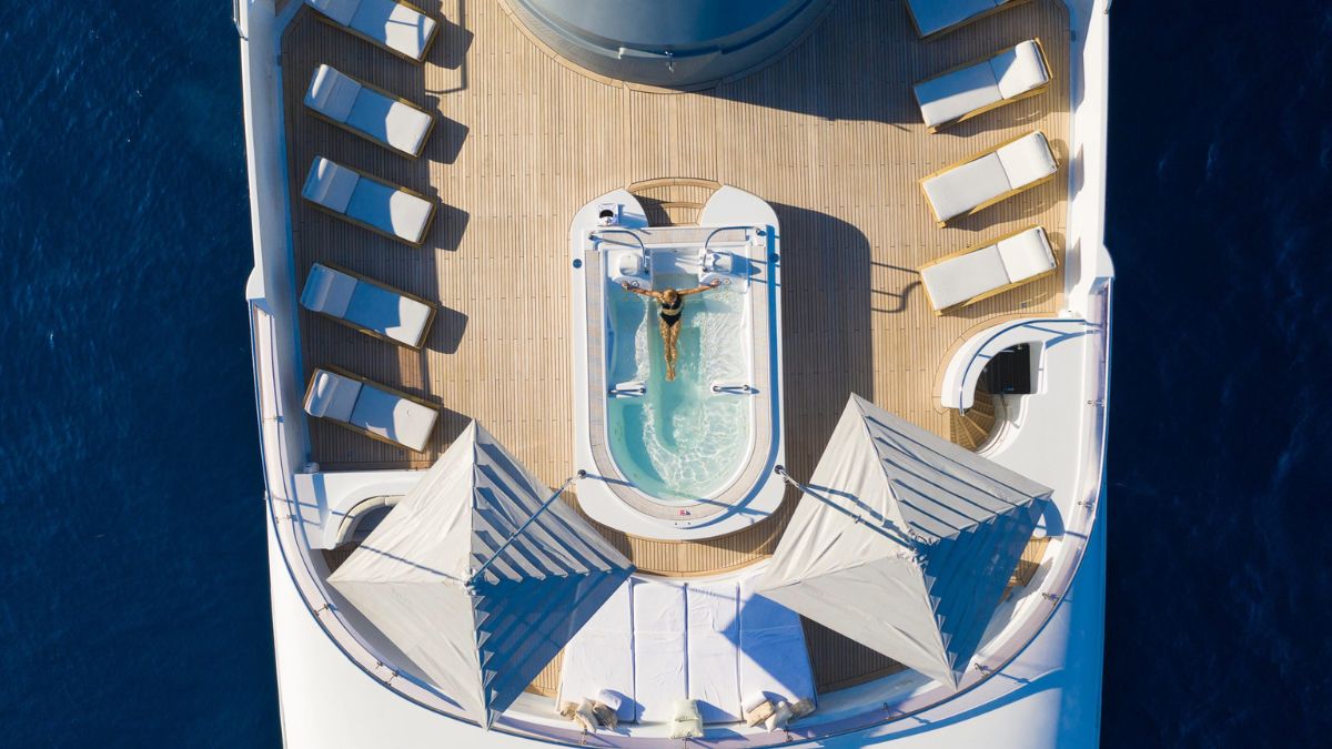 Aerial view of a luxury yacht deck featuring a woman relaxing in a hot tub, surrounded by sun loungers and two large cabanas. The blue ocean surrounds the vessel.