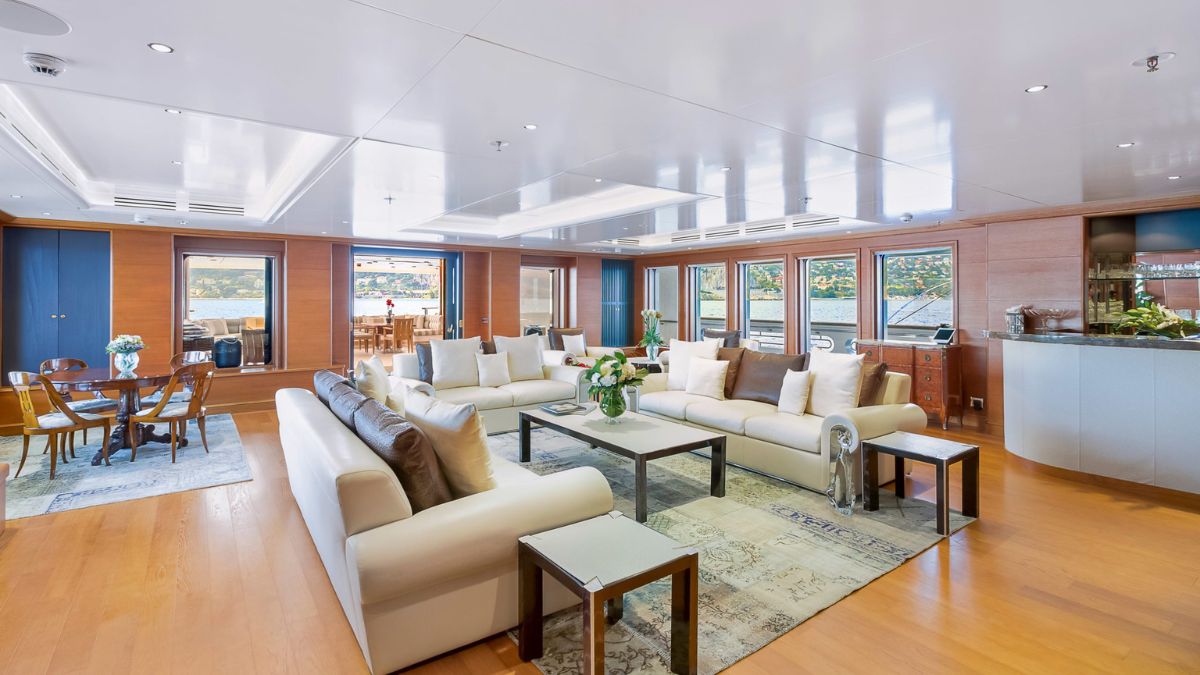Spacious yacht interior featuring a modern, elegantly furnished living area with white sofas, a coffee table, and large windows offering a waterfront view. A dining area is visible in the background, along with a sleek bar. Stylish decor includes light wood accents and vibrant floral arrangements.