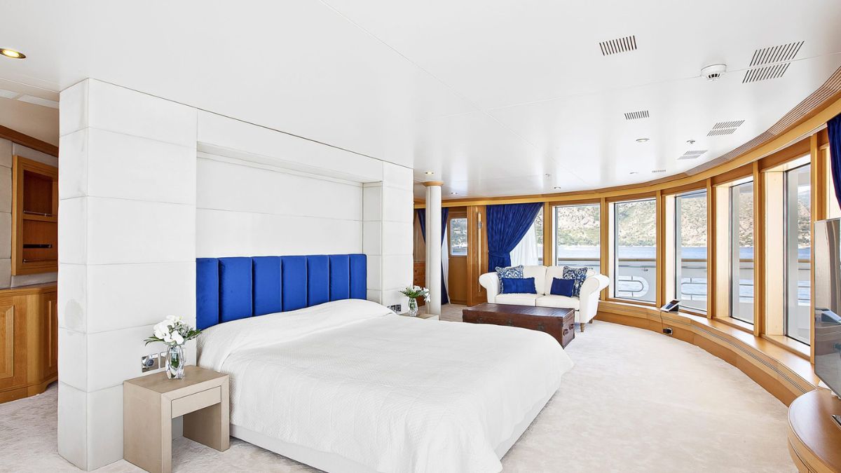 A spacious and modern bedroom featuring a large bed with a white cover, accented by a blue headboard. Large windows line the room, offering a view of the outside, and a cozy seating area with a white couch and decorative pillows is present. The decor is bright and airy, with light-colored walls and plush carpeting.