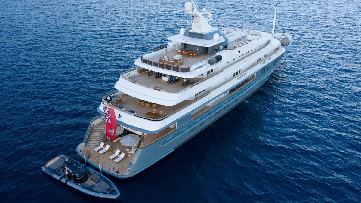 A luxurious yacht anchored in deep blue waters, showcasing multiple decks with sun loungers and a tender alongside. The yacht features a sleek exterior with a flag raised at the stern.