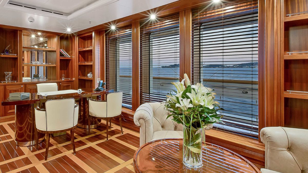 Luxurious office space with wooden panels and polished floors, featuring a glass vase filled with white lilies on a round table. Large windows provide a view of the water, and a desk with elegant chairs is positioned nearby. Soft lighting enhances the sophisticated ambiance.