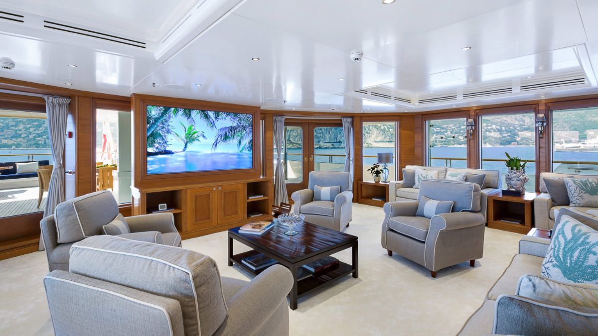 Luxurious yacht interior featuring a spacious lounge with comfortable seating, large windows showcasing scenic views, and a flat-screen TV displaying a tropical scene.