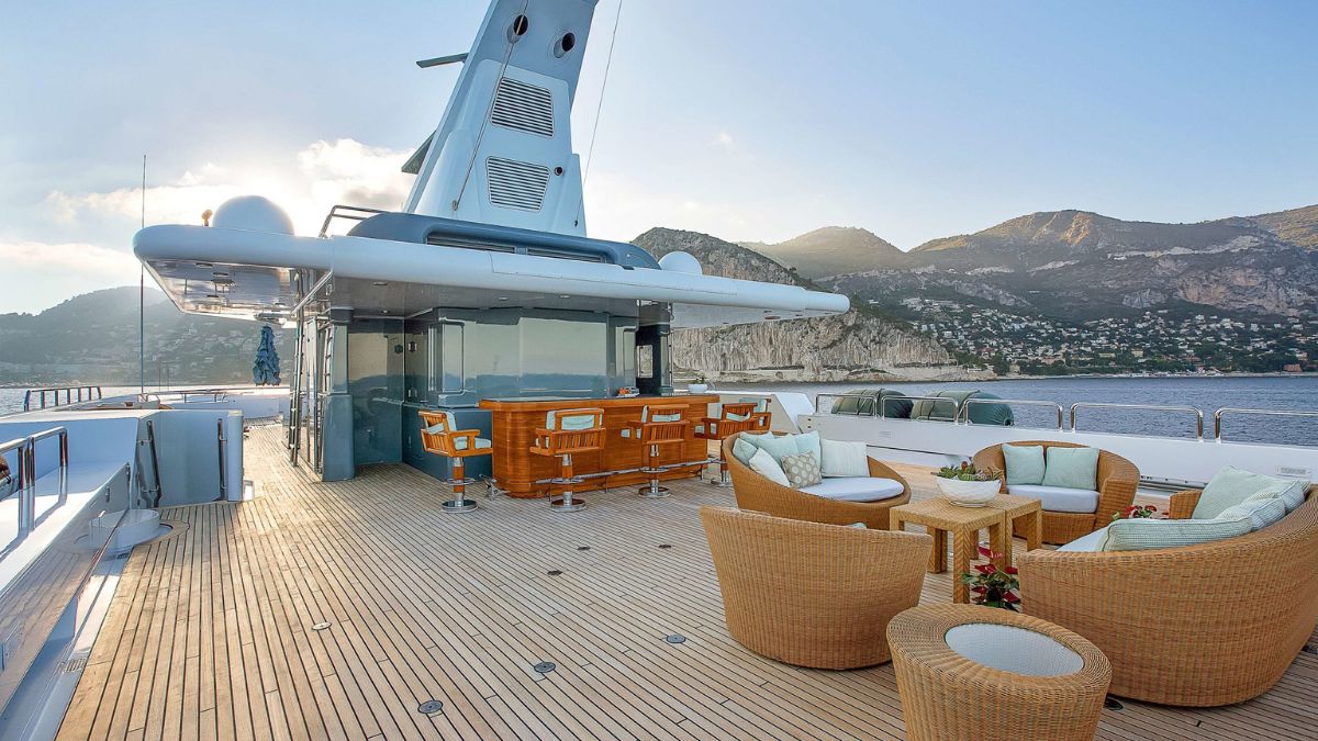 A luxurious yacht deck featuring stylish wicker seating and a bar area with stunning views of mountains and a coastline in the background.