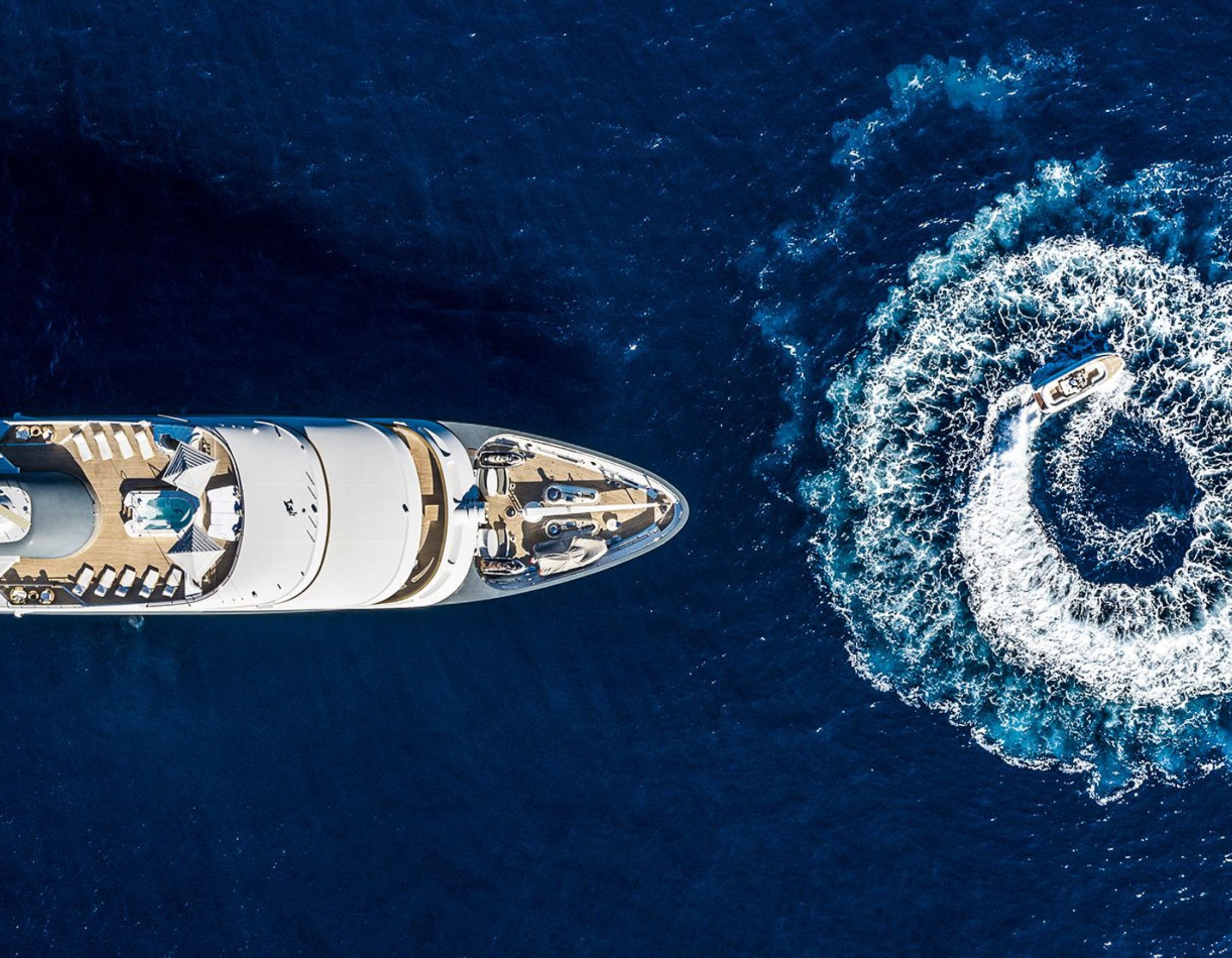 An aerial view of a large luxury yacht anchored in deep blue water, with a small boat creating a circular wake nearby.