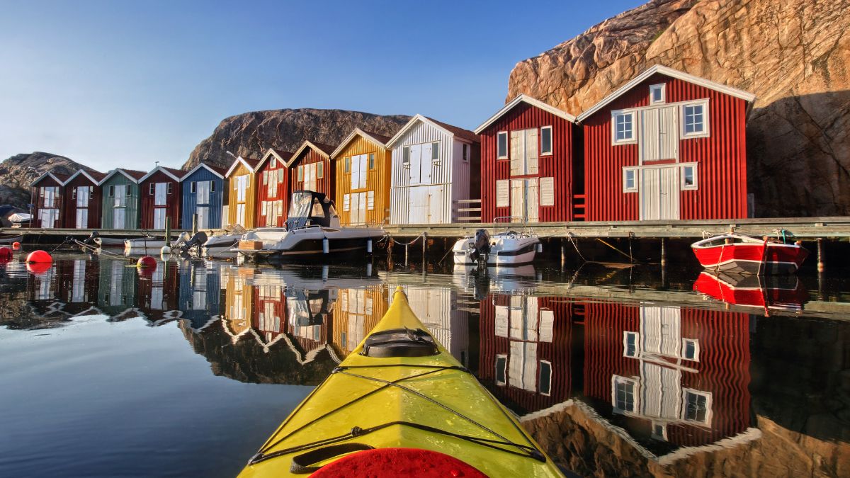 Colorful wooden cabins line a dock by a calm, reflective body of water, with kayaks and boats moored nearby. Rocky cliffs rise in the background under a clear blue sky.