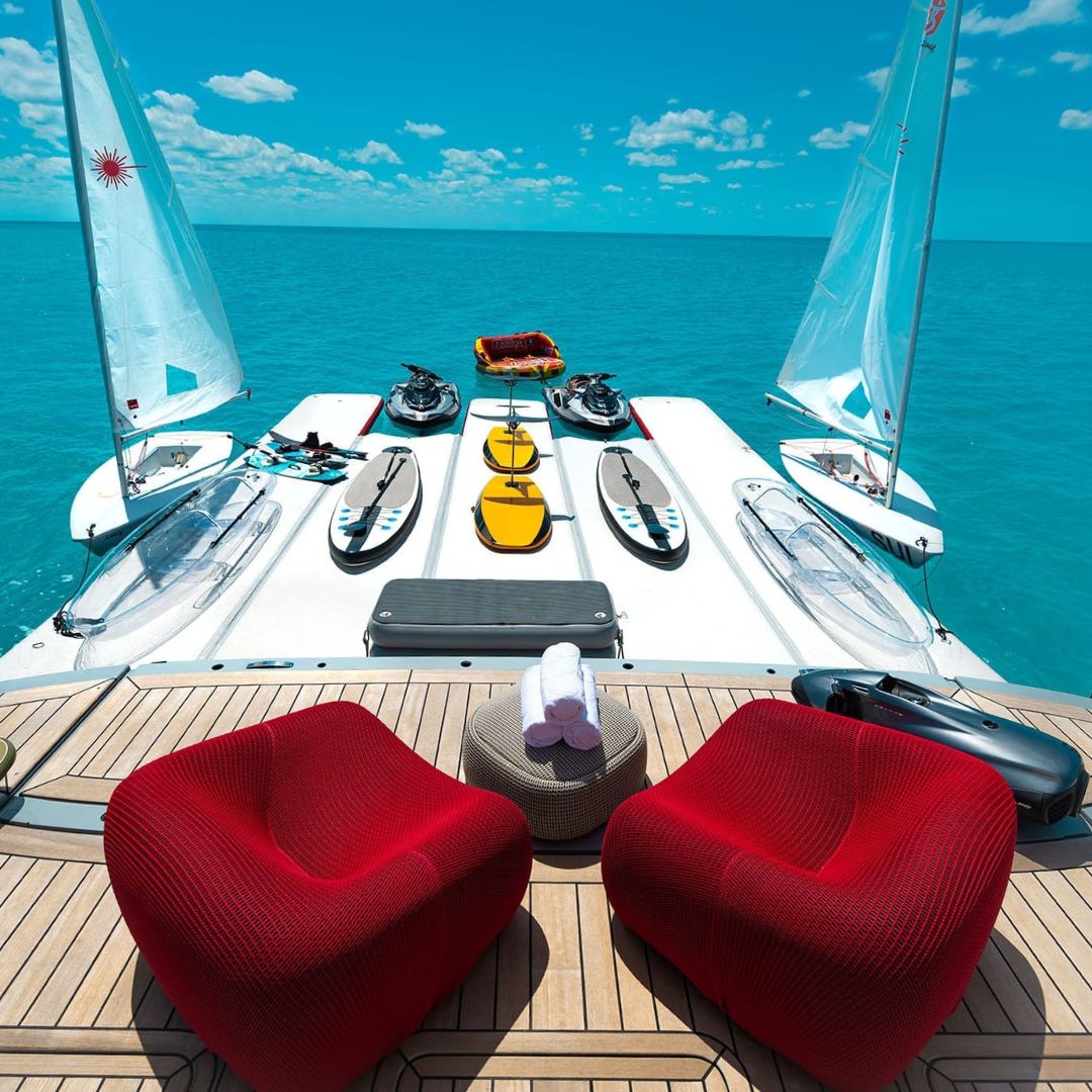 A luxurious yacht deck with vibrant red chairs overlooking a turquoise sea. Various kayaks and sailboats are moored alongside, set against a clear blue sky with fluffy clouds.