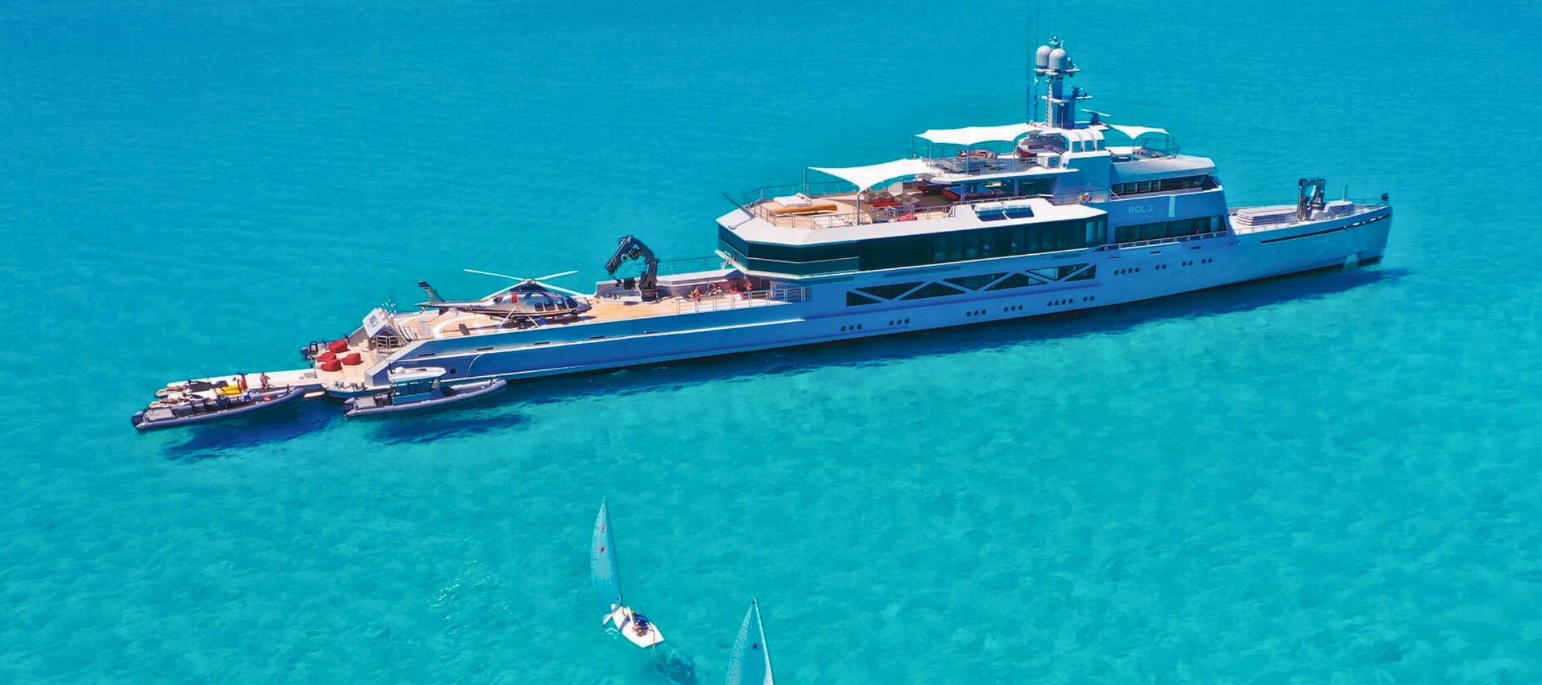 A large luxury yacht anchored in vibrant turquoise waters, with small dinghies sailing nearby. The yacht features multiple decks, a helicopter on the top, and various water sports equipment visible.