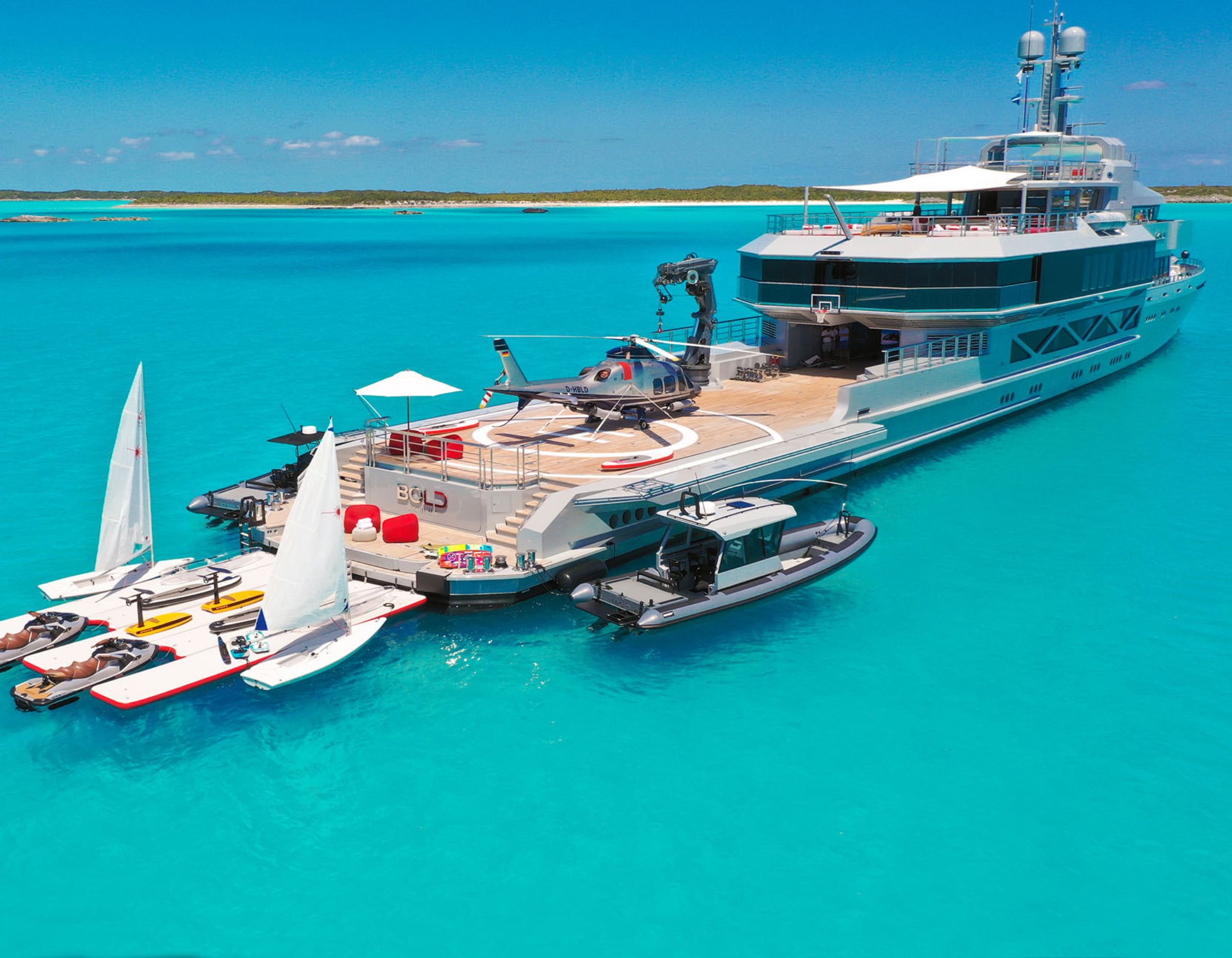 A luxurious yacht anchored in clear turquoise waters, featuring a helicopter on the deck, small sailboats at the rear, and a sleek tender boat. A beach is visible in the background under a bright blue sky.