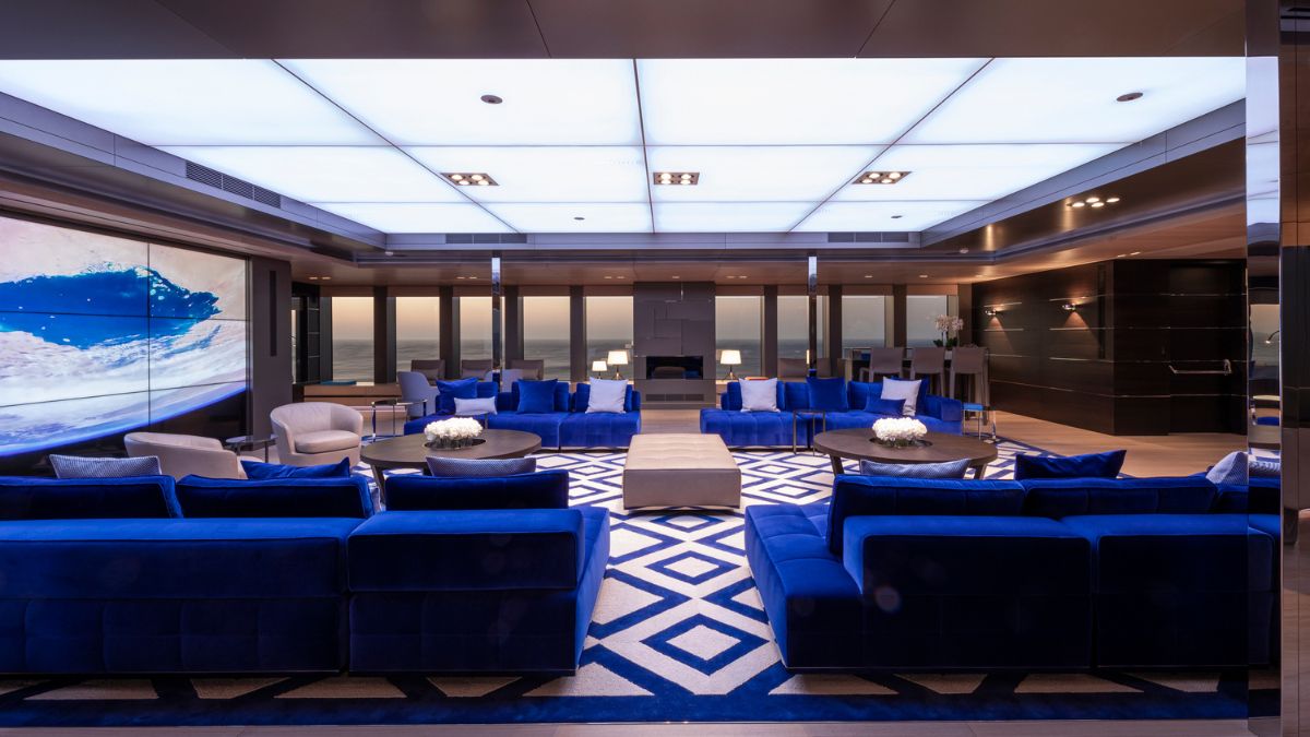 A luxurious modern lounge featuring deep blue velvet seating, a light-colored coffee table, and elegant decor. Large windows provide a view of the ocean, and a stylish rug adds to the ambiance. The ceiling features a sleek, illuminated design.