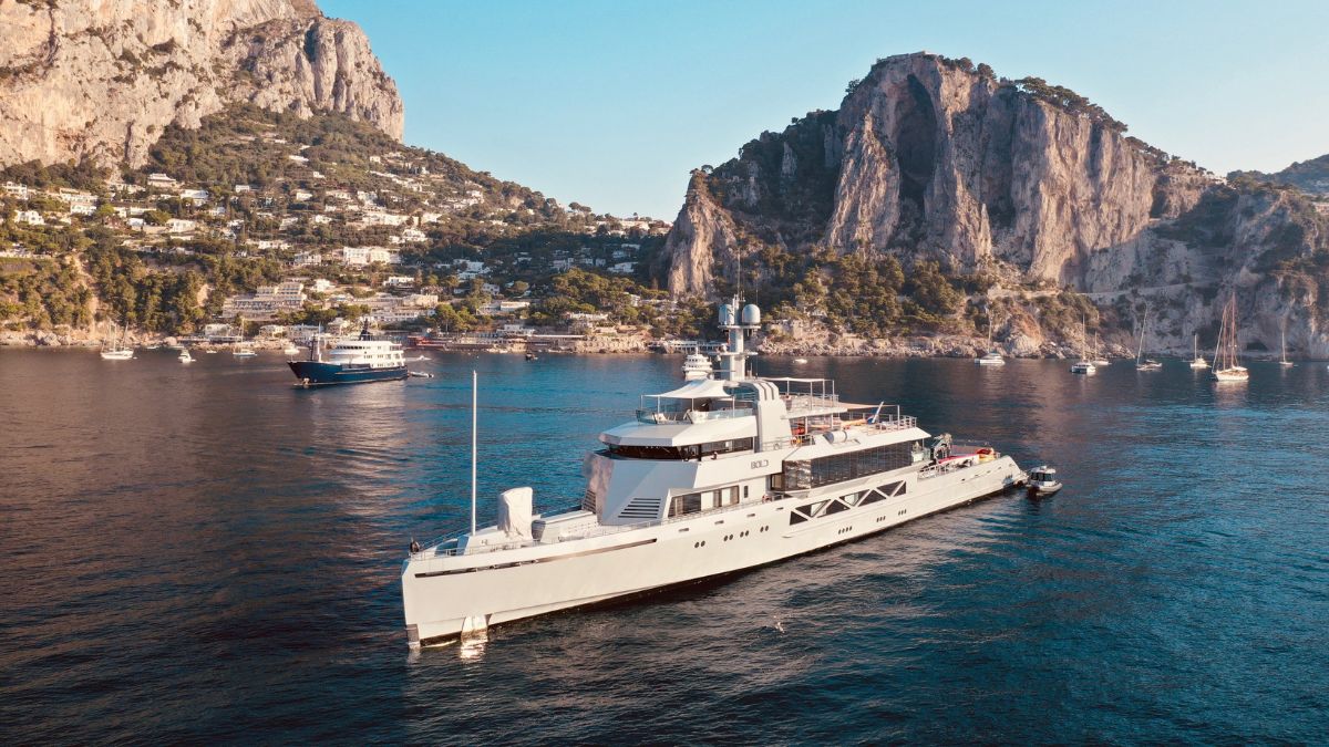A luxury yacht anchored in calm blue waters near a rocky coastline. The background features steep cliffs and lush greenery, with several smaller boats nearby.