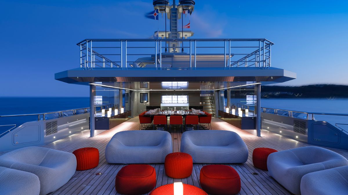 A luxurious yacht deck at dusk, featuring a modern dining area with a long table and red chairs, surrounded by stylish lounge seating. Soft lighting highlights the space against a backdrop of a serene ocean.