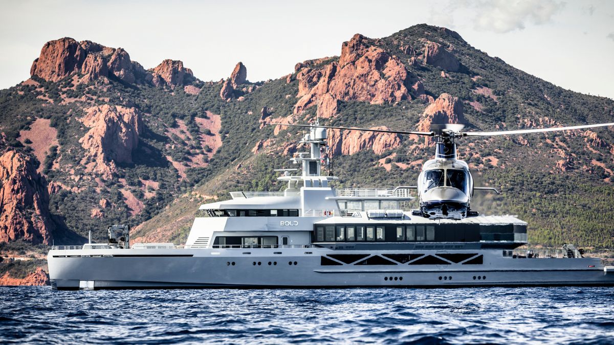 A sleek, modern yacht named Bold is anchored in calm waters with a helicopter hovering above it. Dramatic, rocky mountains rise in the background, framed by lush greenery.