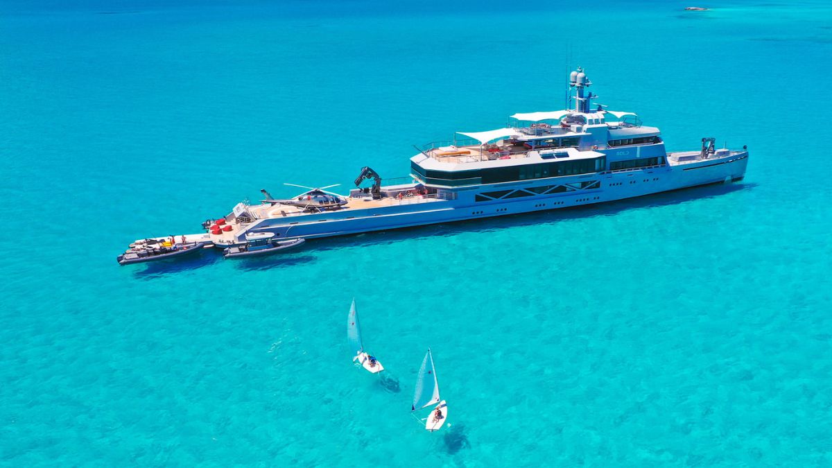 A large luxury yacht anchored in clear turquoise waters, with two smaller sailboats nearby. The yacht features multiple decks and a helipad on top.