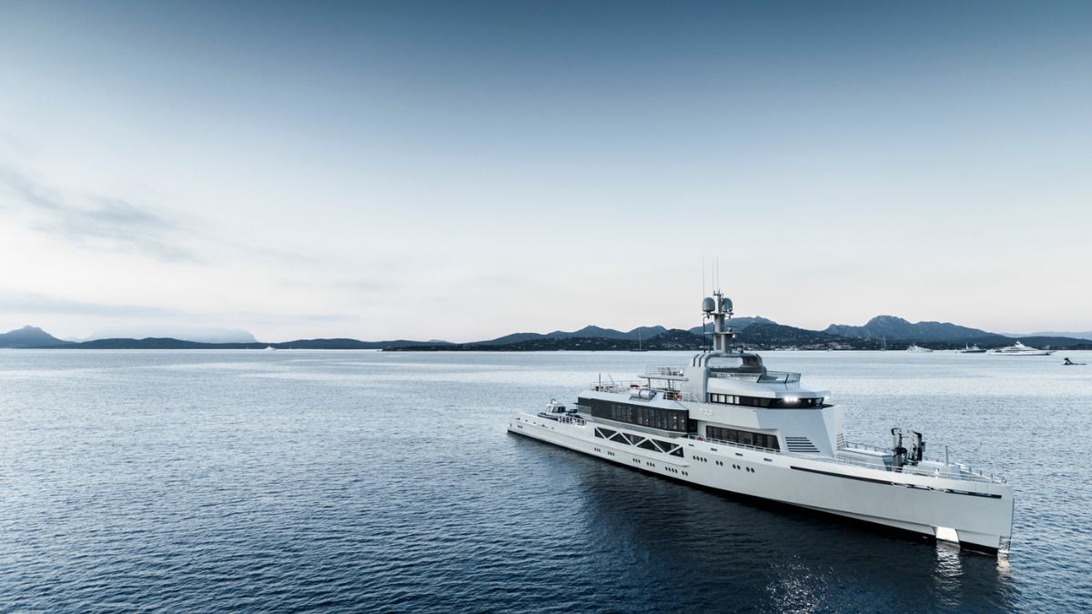 A sleek superyacht anchored in calm waters, surrounded by distant hills and a clear sky. The yacht features modern design elements, with a prominent helipad and luxurious deck spaces.