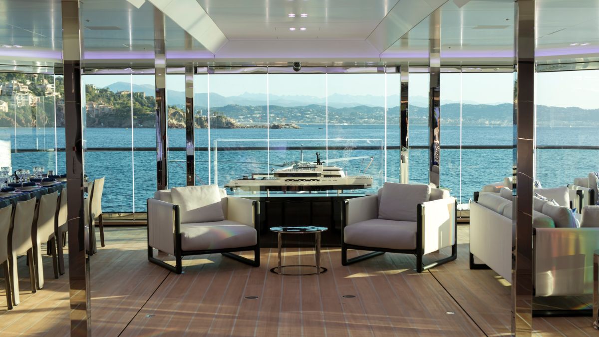 Luxurious yacht interior featuring modern furniture with two white armchairs and a glass table, overlooking a stunning ocean view through large windows. The space is elegantly decorated with a contemporary design, showcasing a sleek dining area in the background.