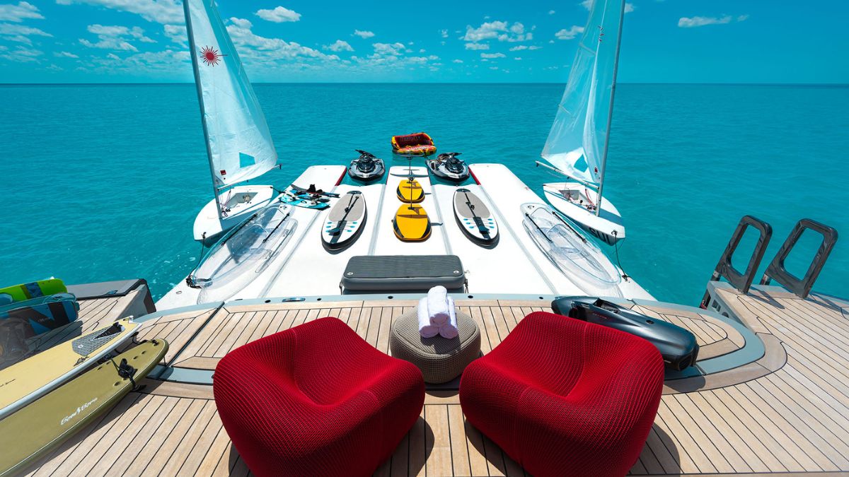 A luxurious catamaran deck overlooking a turquoise sea, featuring two vibrant red chairs and various water sports equipment, including kayaks and paddleboards, ready for adventure.
