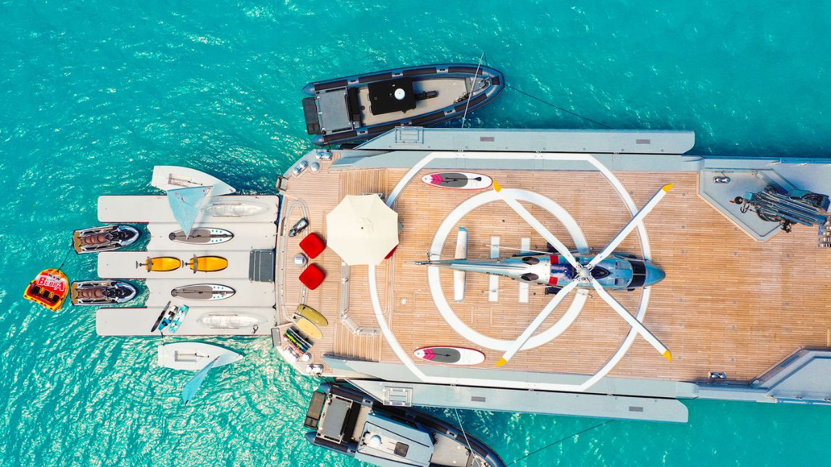 A luxurious yacht docked in turquoise waters, featuring a helipad at its centre. Surrounding the helipad are various boats and jet skis, with a parasol and seating area visible on the deck.