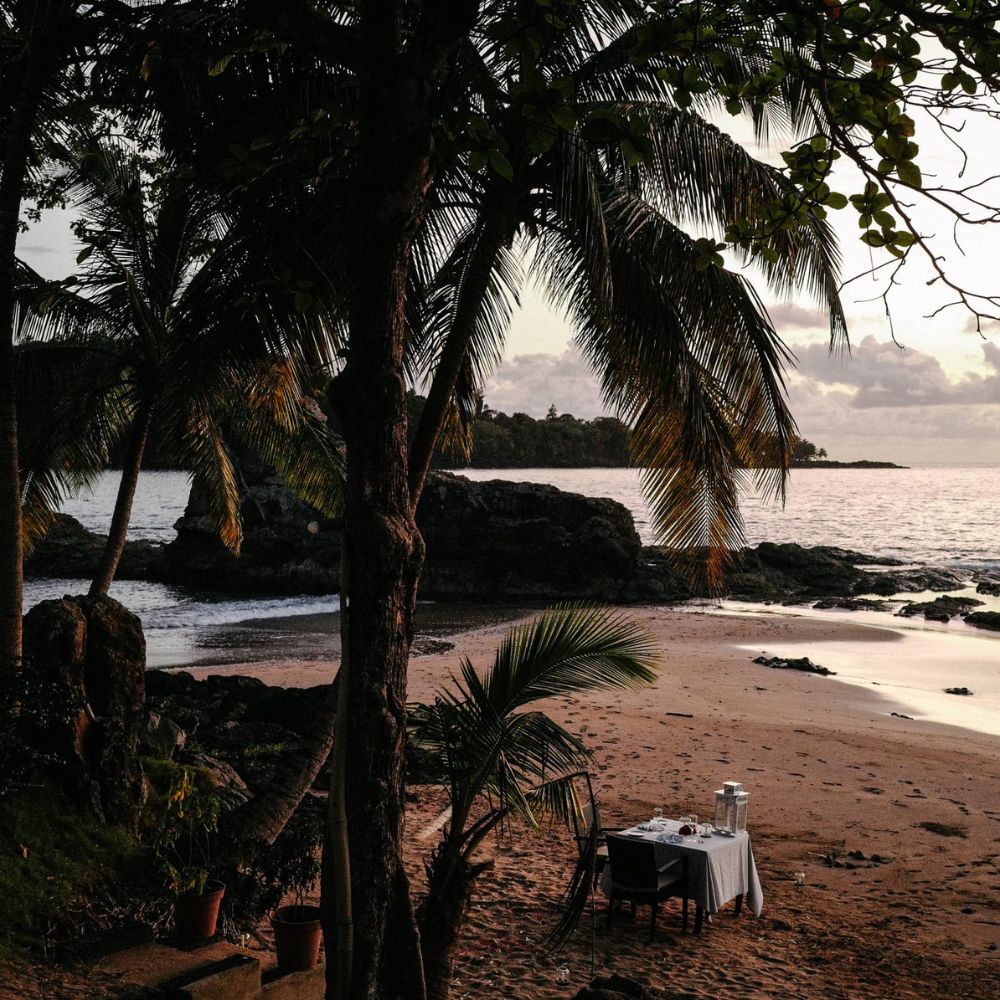 A secluded beach at sunset featuring a table set for two, surrounded by palm trees and rocky outcrops. The soft waves gently lap at the shore while the sky displays warm hues of orange and pink.