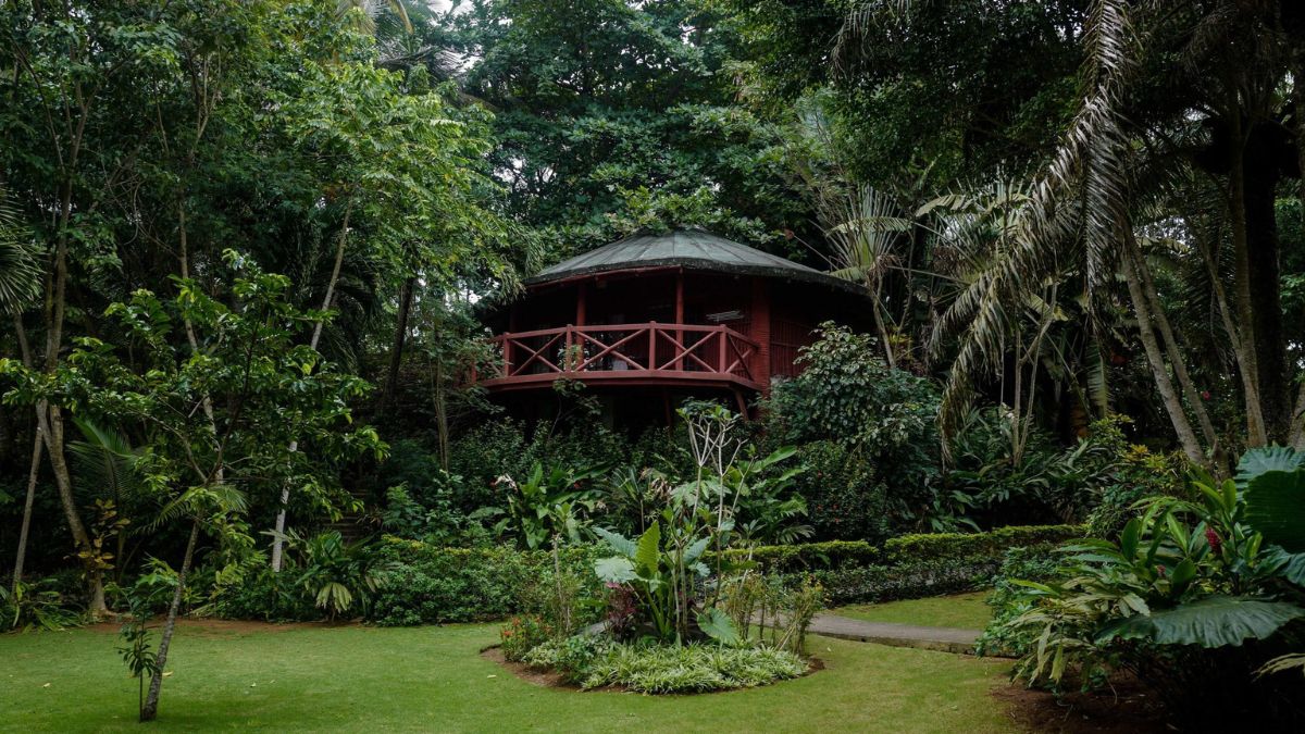 A red cottage with a circular design surrounded by lush greenery and tropical plants. A well-maintained lawn is in the foreground with neatly arranged flower beds and pathways leading to the cottage.