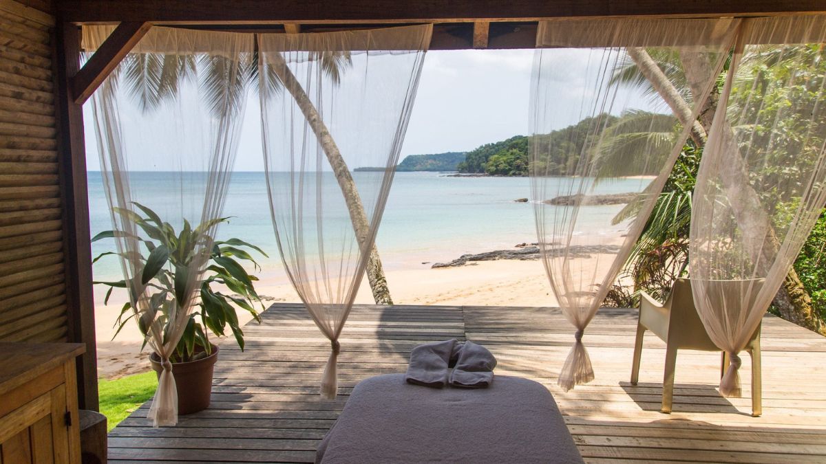 A serene beach view from a wooden terrace, featuring sheer curtains fluttering in the breeze. A massage table is set up in the foreground, surrounded by tropical plants, with a calm sea and sandy shore in the background.