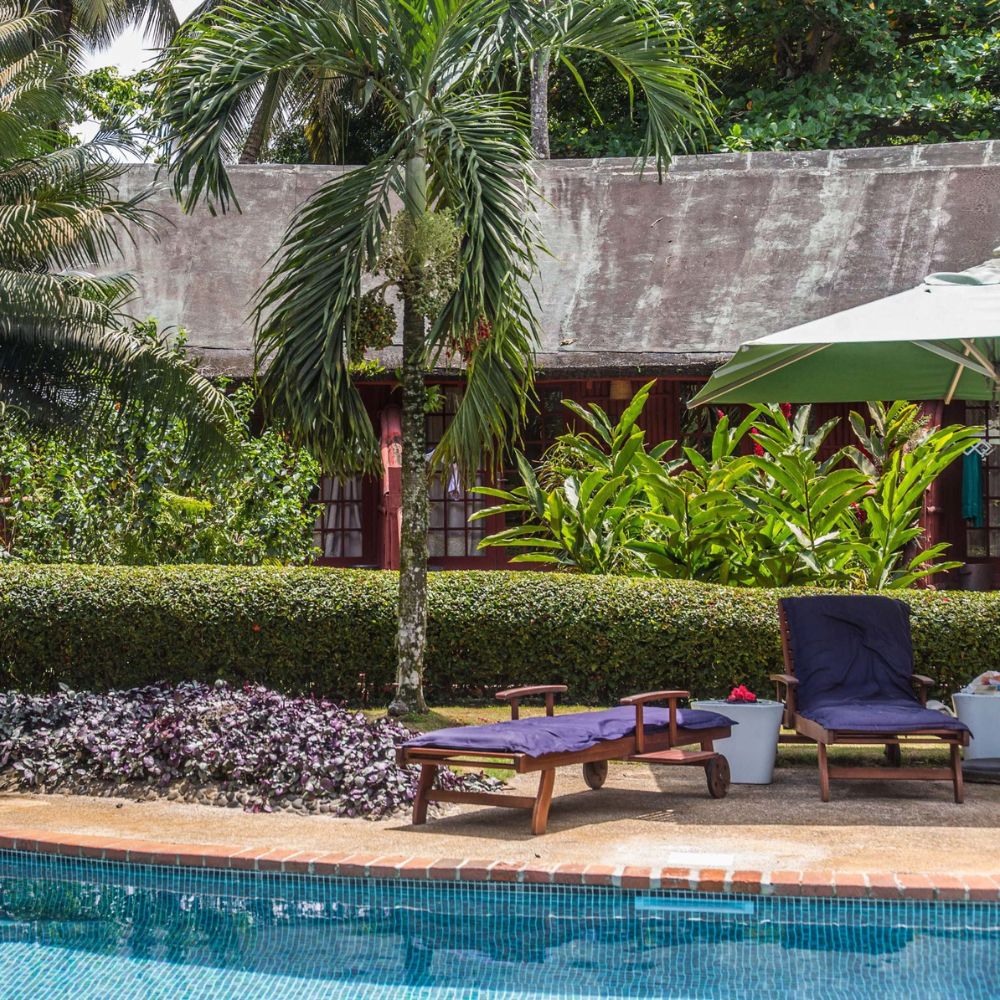 A serene poolside scene featuring two lounge chairs by a clear blue pool. Lush greenery, including palm trees and colorful plants, surrounds the area. In the background, a rustic building with a thatched roof is partially visible.