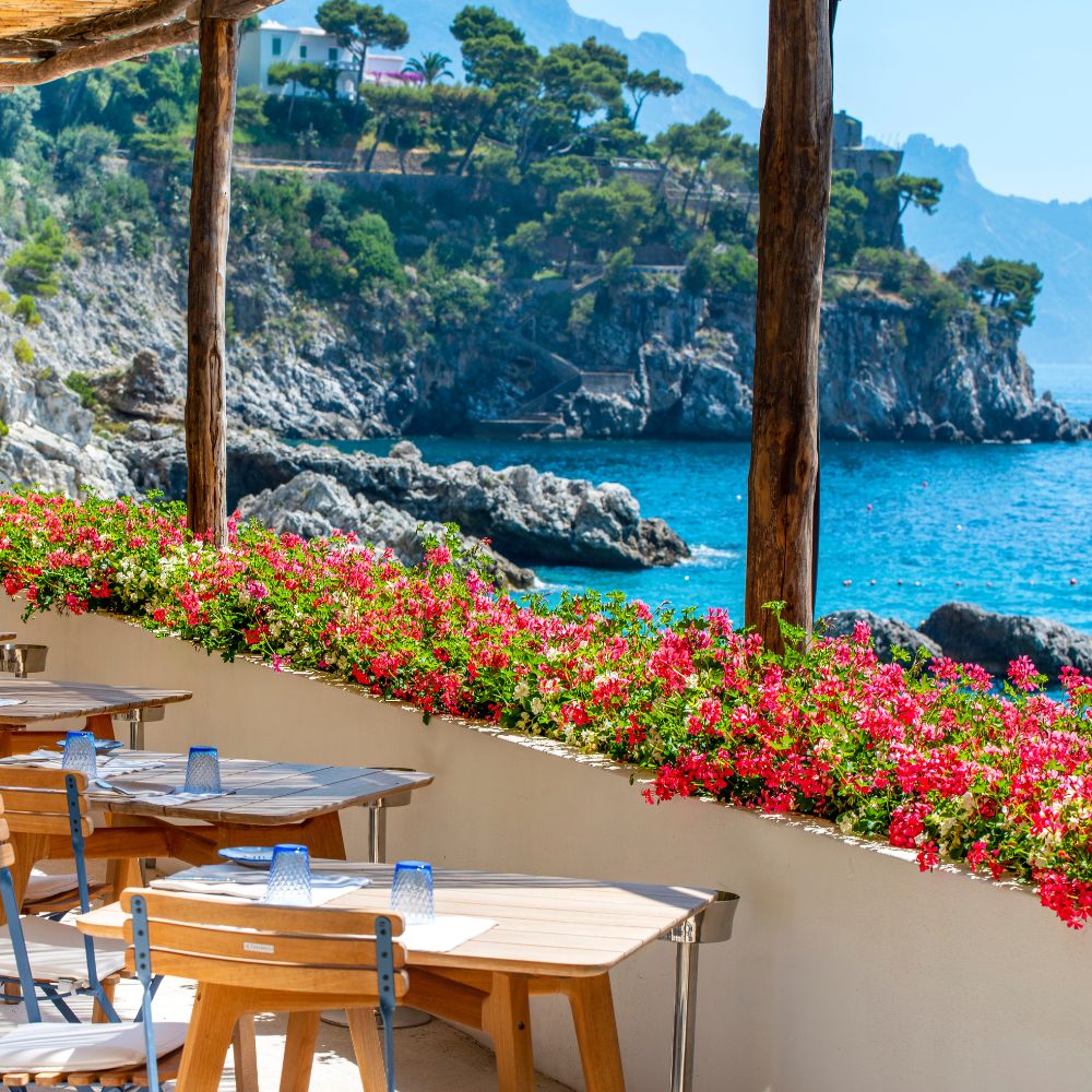 Seaside dining area with wooden tables and blue glasses, adorned with vibrant pink and red flowers. A picturesque rocky coastline and clear blue water are visible in the background, framed by lush greenery and mountains.