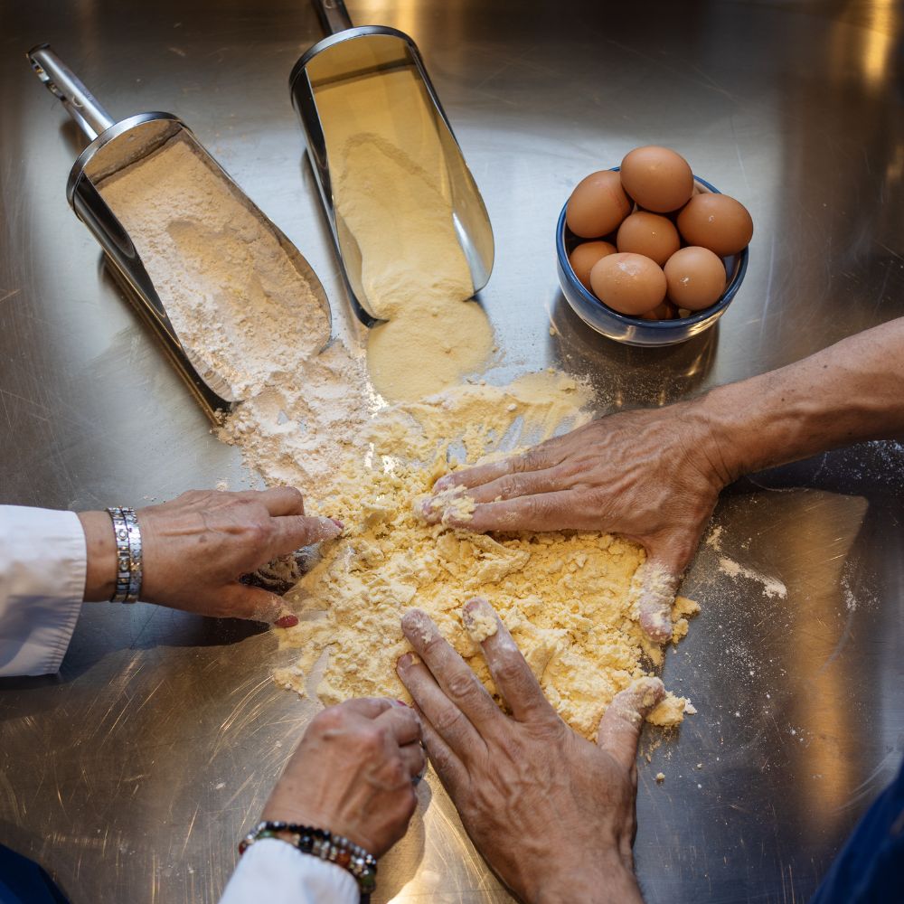 Hands kneading dough on a stainless steel surface, surrounded by flour and a bowl of eggs. Two scoops of flour and cornmeal are nearby.