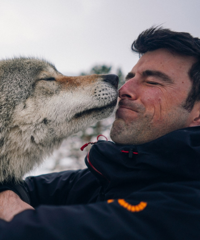A man and a wolf share a tender moment, their faces close together as the wolf nuzzles against him. The man appears relaxed and happy, wearing a black jacket. The background is softly blurred, suggesting a natural setting.